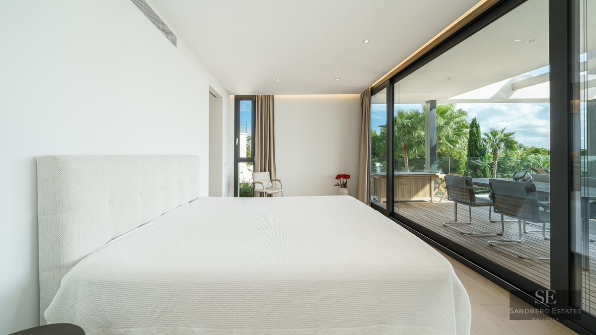 Minimalist white master bedroom with floor-to-ceiling glass doors opening onto a sunny wooden terrace with palm trees.