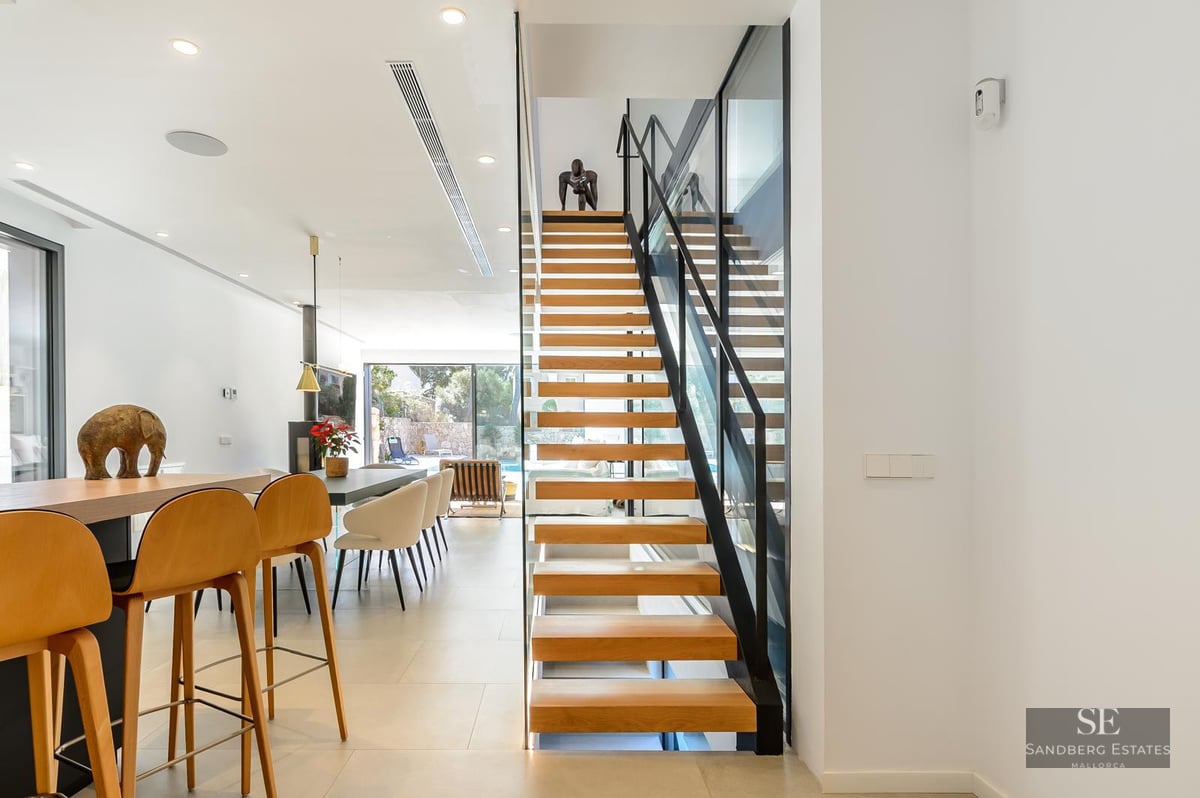 A modern interior showing a wooden floating staircase with glass railings and a bright living area.