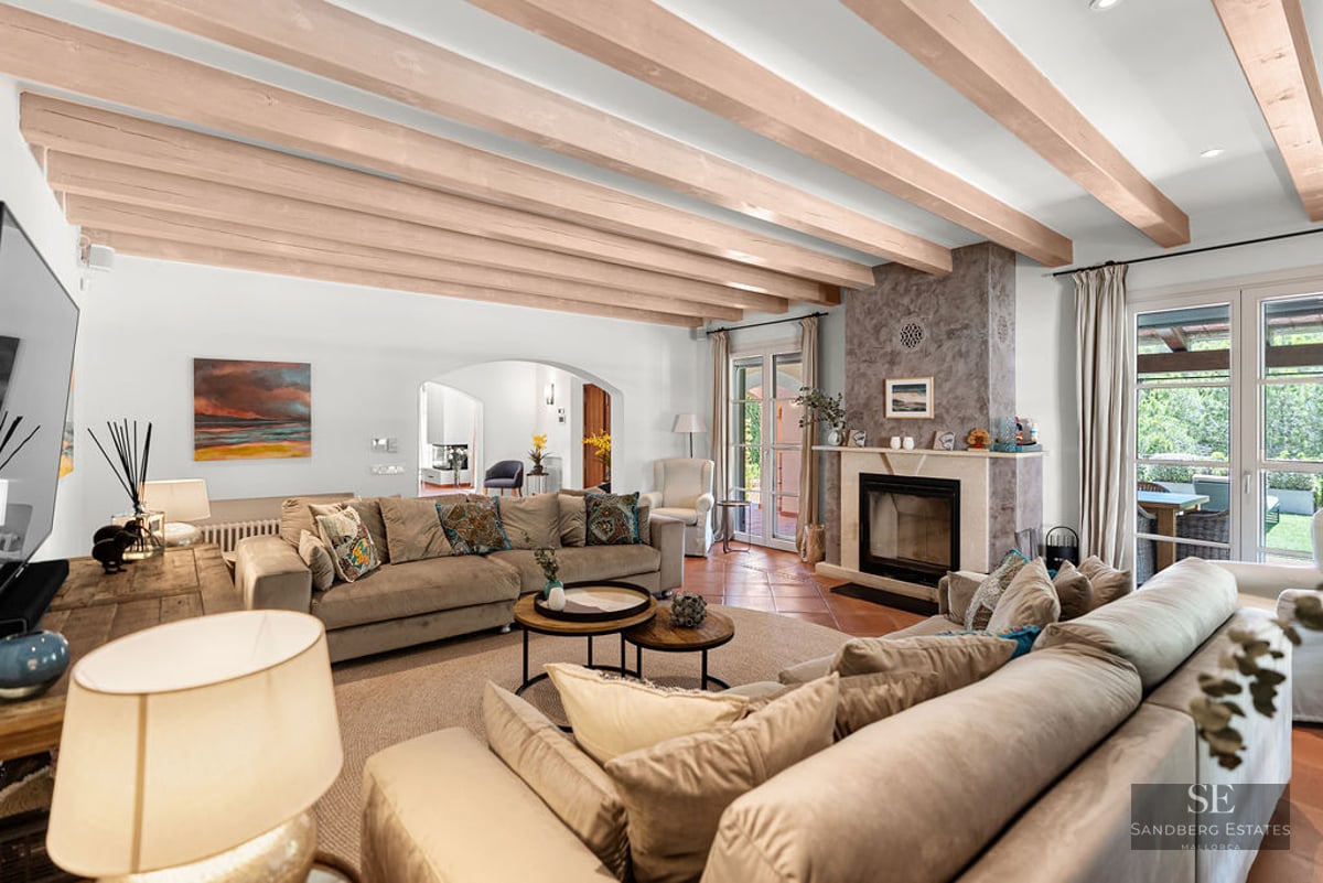 Spacious living room with beige sofas, stone fireplace, wooden ceiling beams, and terrace access.