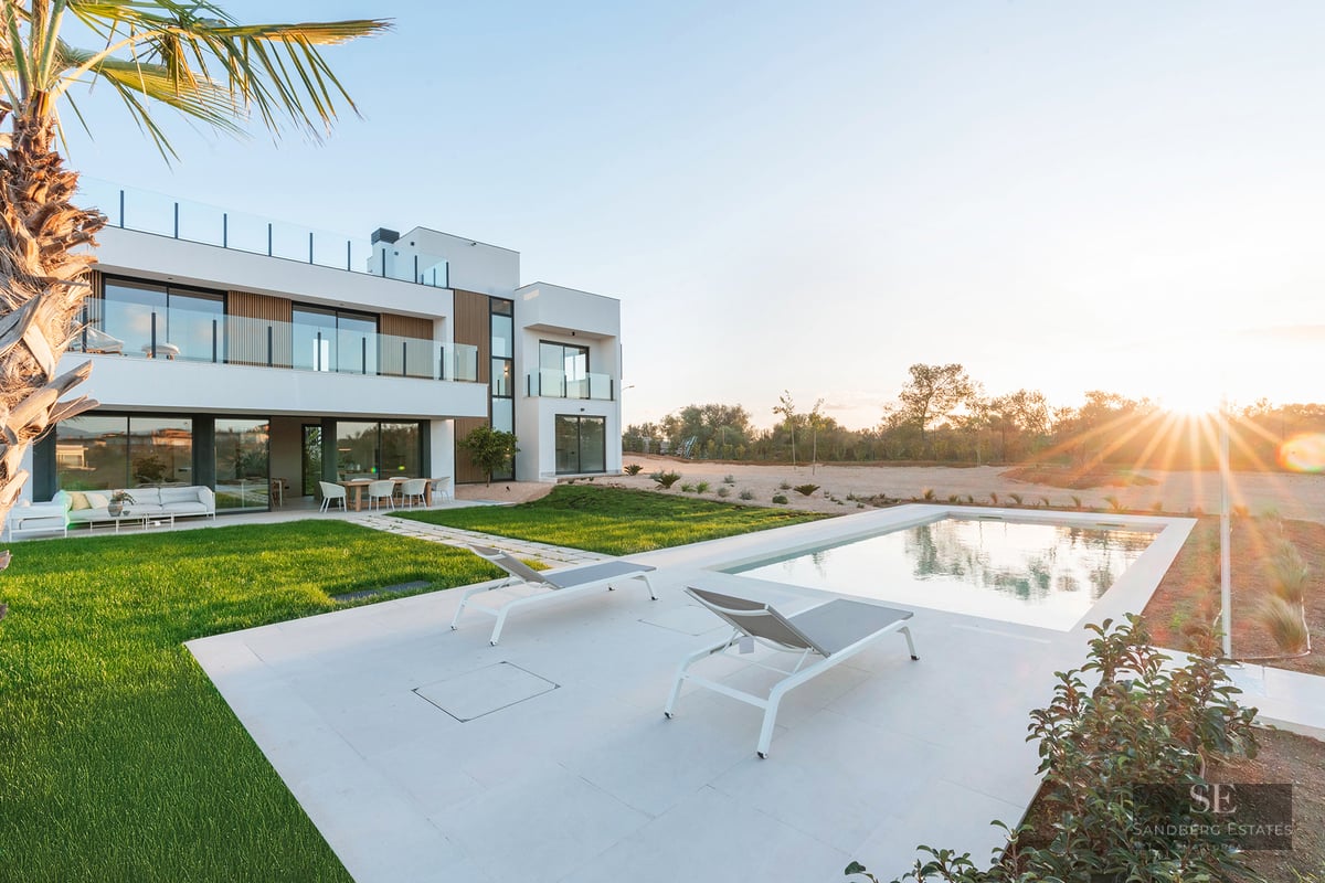Modern white two-story villa with a swimming pool, sun loungers, and green lawn under a golden sunset sky.