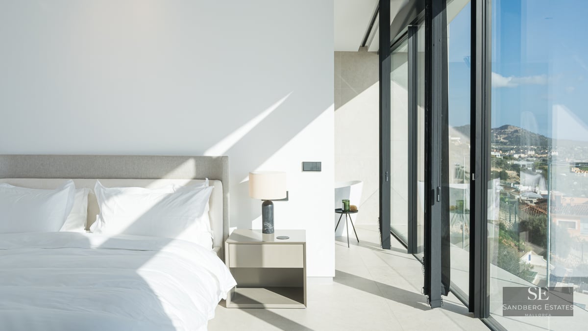 Bright minimalist master bedroom with floor-to-ceiling glass windows and white linens under natural sunlight.