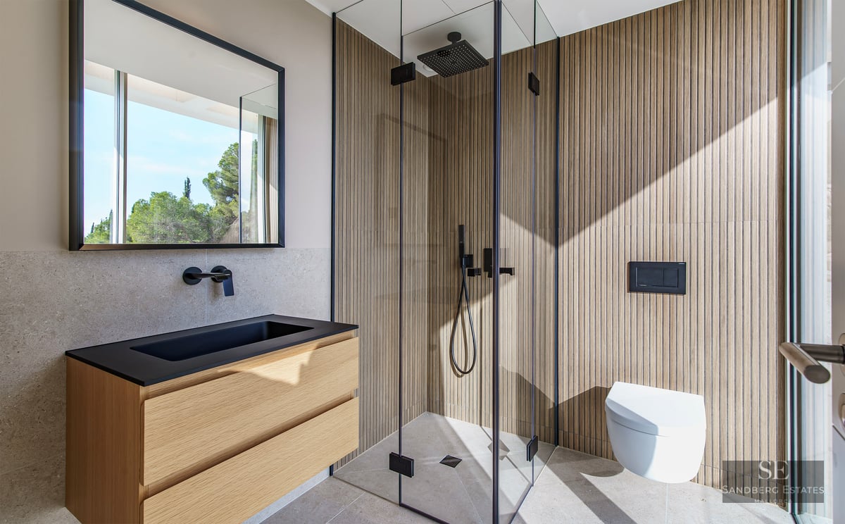 Modern bathroom with wood-slat walls, glass shower, black vanity, and wall-mounted toilet under bright natural light.