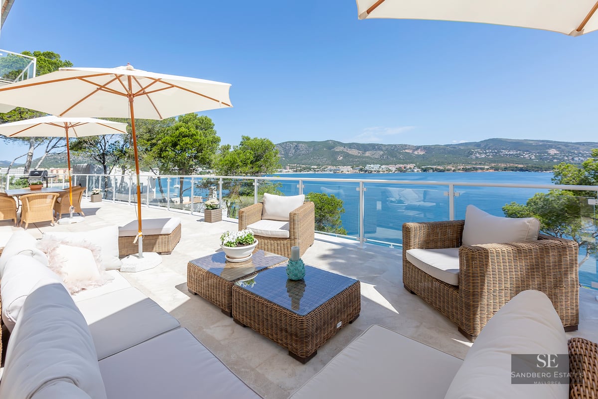 Sunlit terrace with wicker lounge furniture, white umbrellas, and glass railings overlooking a blue sea and mountains.