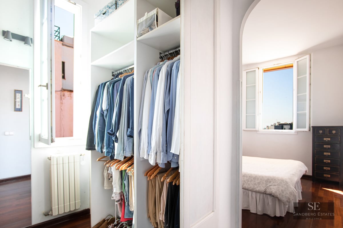 Open white closet with clothes hanging next to a window, looking into a sunlit bedroom.