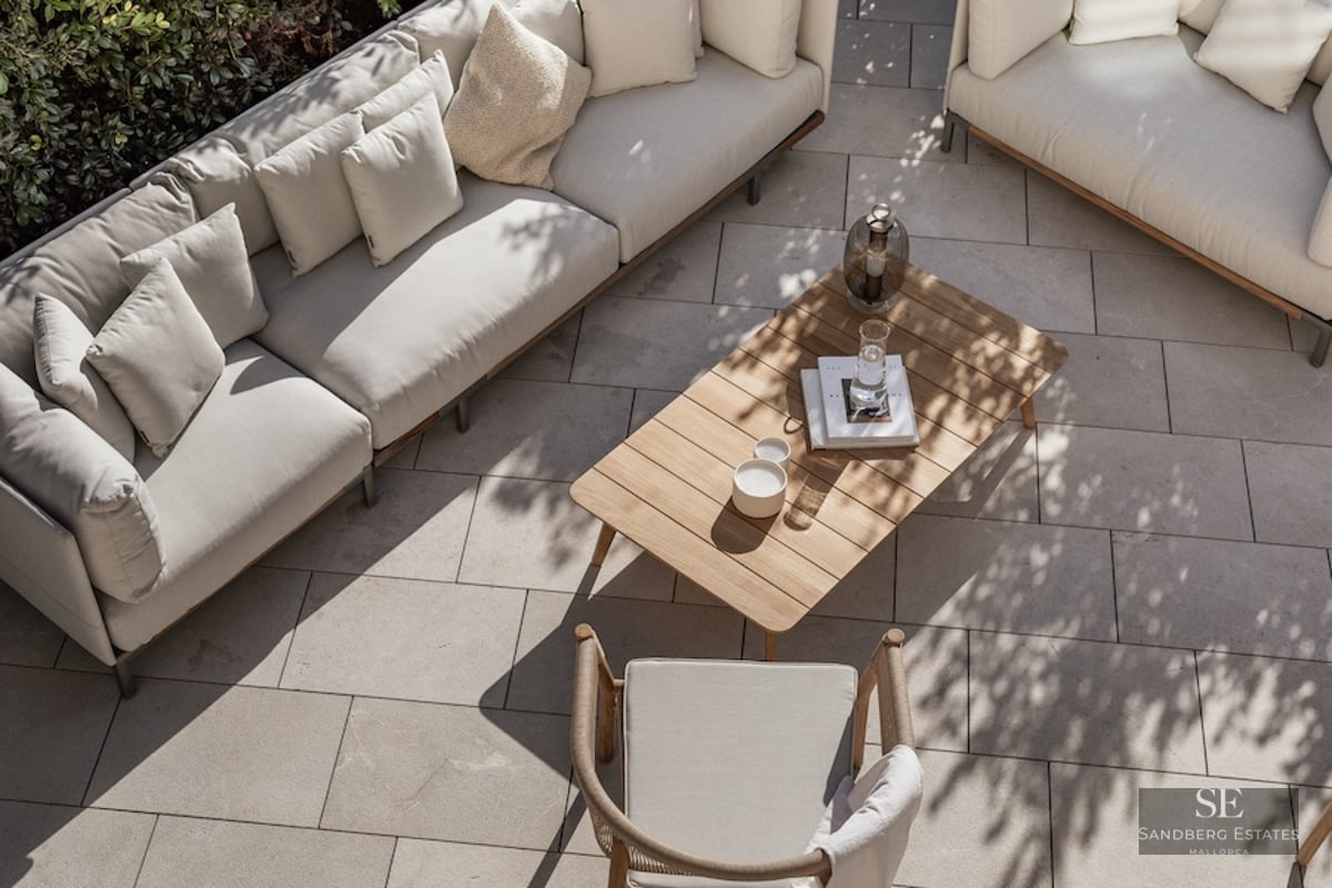 High-angle view of a luxury terrace with beige sofas, a wooden coffee table, and stone paving in dappled sunlight.