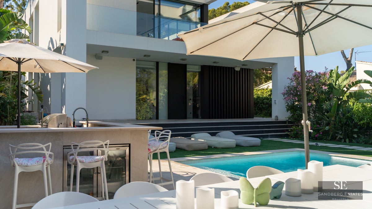 Modern swimming pool with lounge bean bags, outdoor kitchen, and white umbrellas next to a contemporary villa.