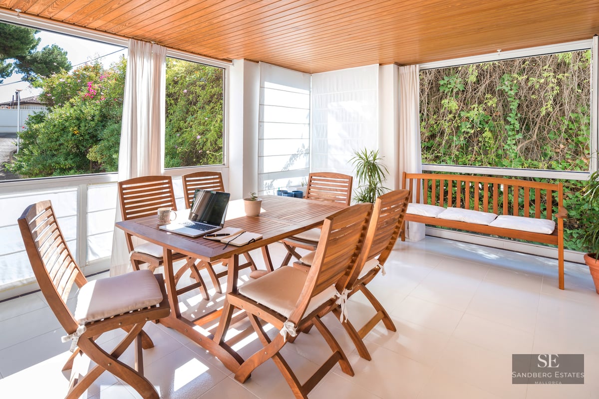 A sunlit terrace room with a wooden dining table, chairs, and bench, overlooking lush green garden foliage.