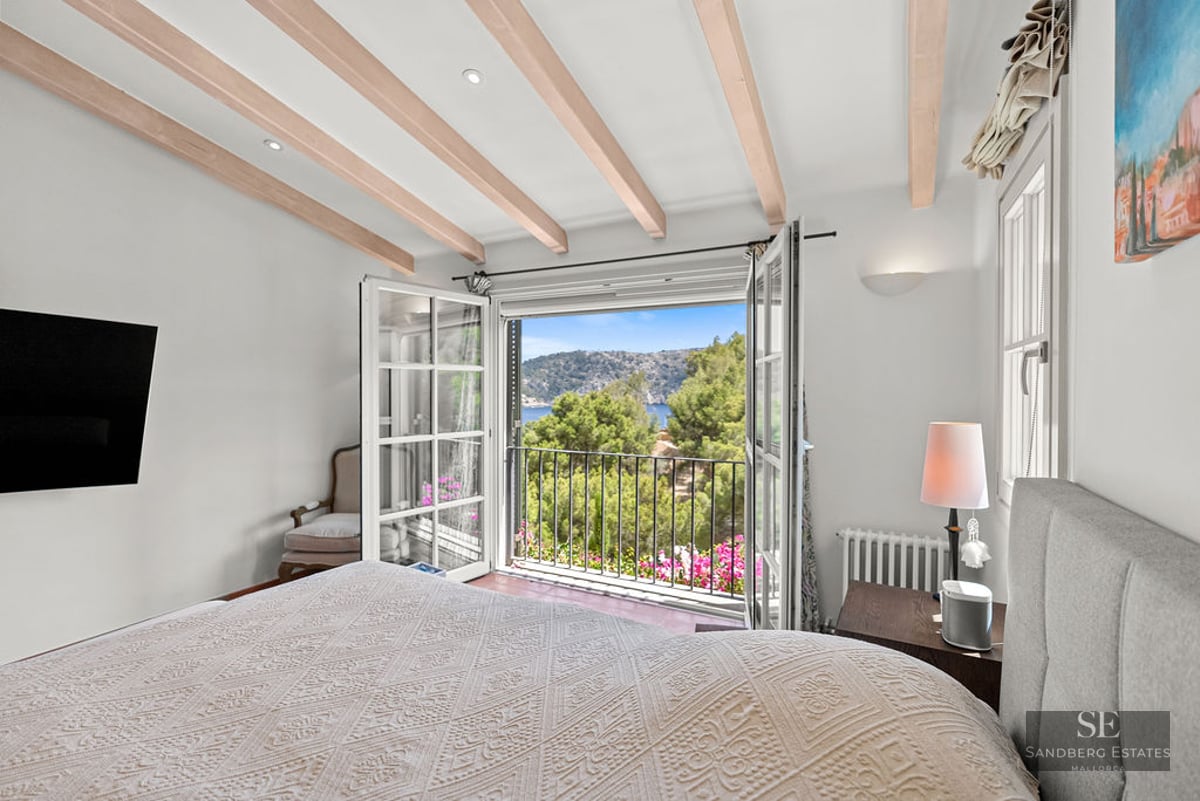 Bright bedroom with wooden ceiling beams and open balcony doors revealing a lush green hillside and sea view.