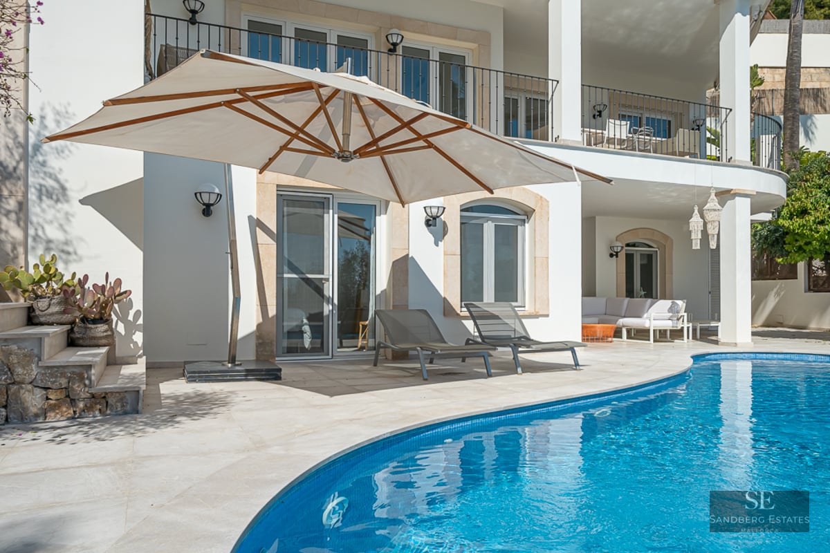 Piscine bleue incurvée avec terrasse en pierre blanche, grand parasol beige et transats devant une villa blanche.