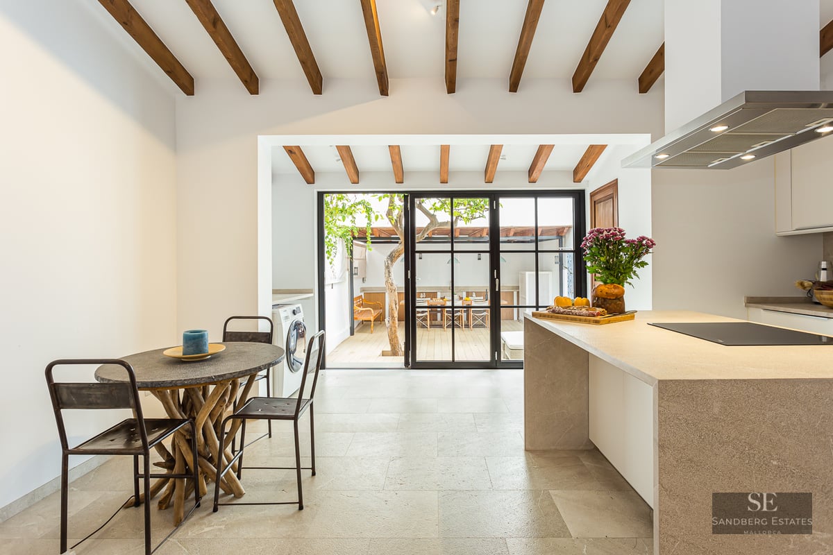 Bright kitchen with wood ceiling beams, stone island, and glass doors opening to a sunny outdoor patio with a tree.