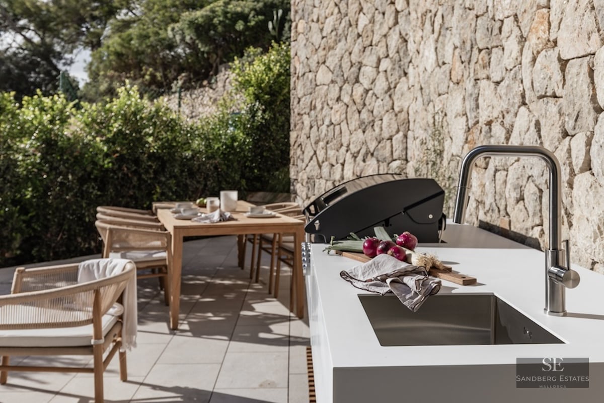 Modern outdoor kitchen with white countertop, sink, and grill next to a stone wall and wooden dining set.