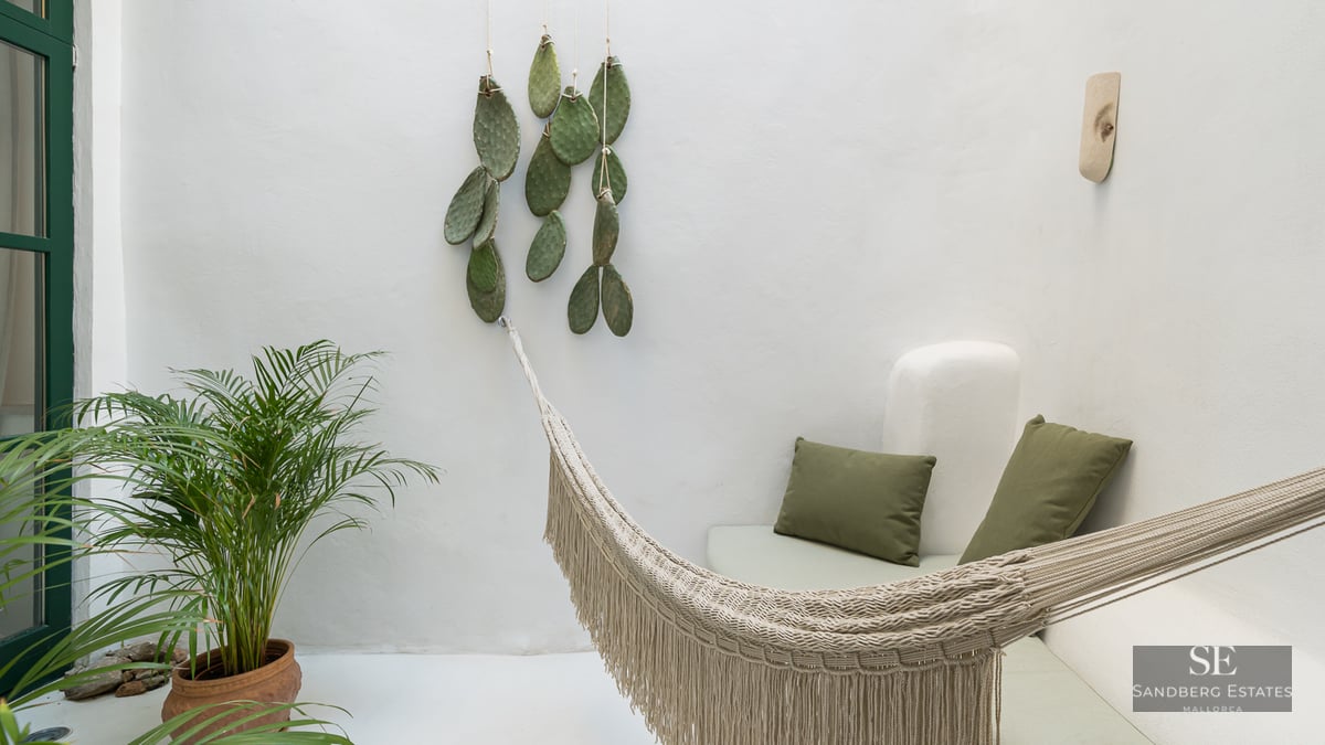 A white-walled outdoor terrace featuring a woven fringed hammock, olive green pillows, and a potted palm plant.
