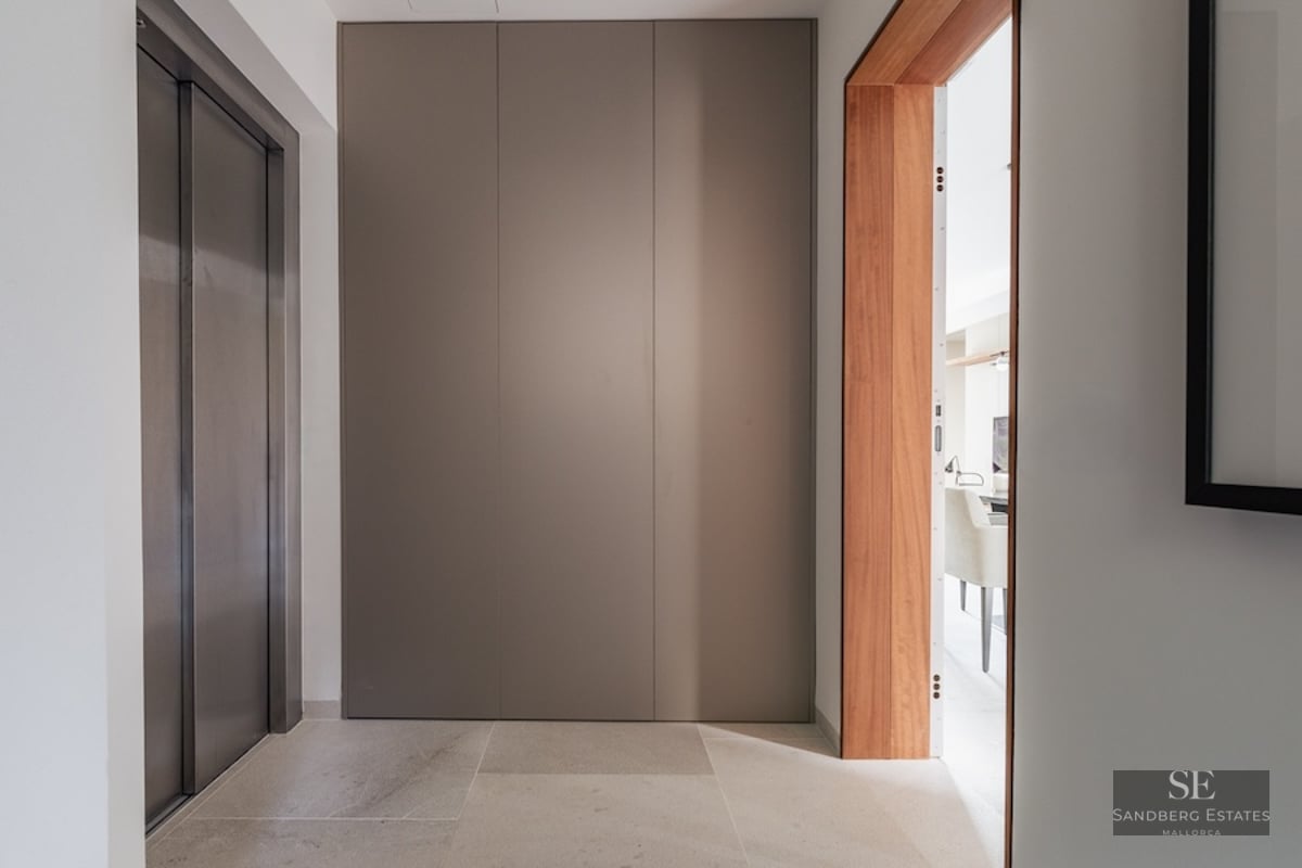 Modern entrance hallway featuring a metallic elevator, taupe panels, and a warm wooden door frame.