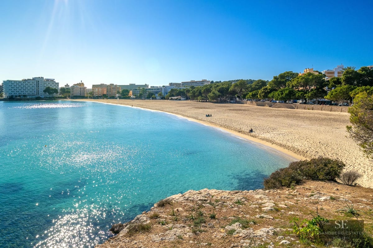 High-angle view of a golden sand beach and sparkling turquoise sea under a bright blue sunlit sky.