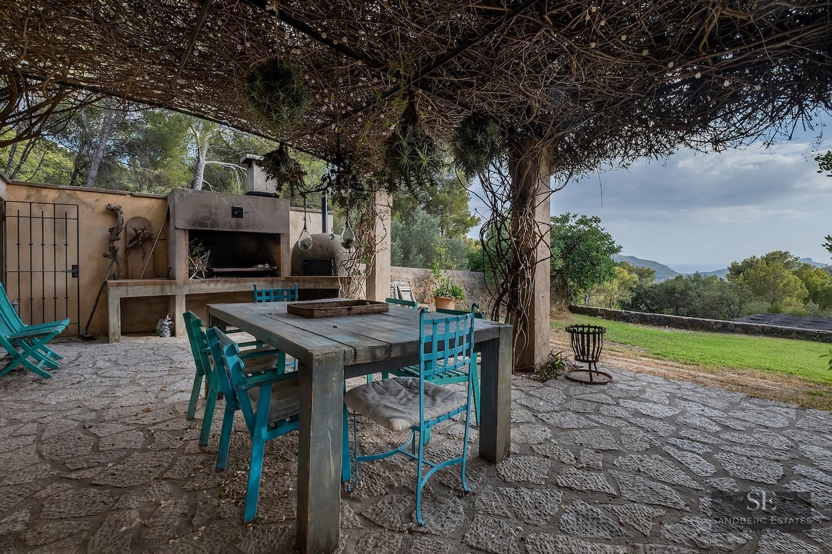 Rustic stone terrace with turquoise chairs, wooden table, and BBQ under a vine-covered pergola overlooking hills.