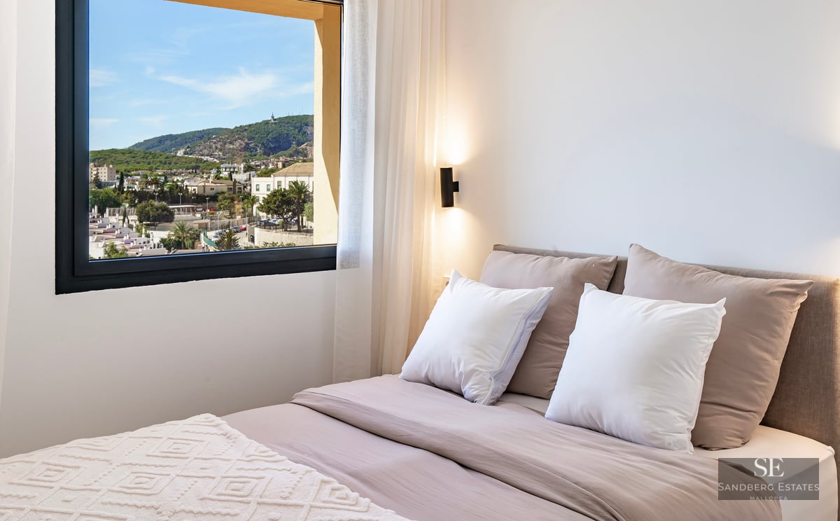 Modern bedroom with neutral-toned bedding and a large window overlooking a coastal town and green hills.