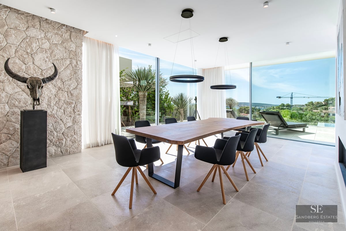 A modern dining room featuring a large wooden table, black chairs, a stone wall with a skull sculpture, and large windows.