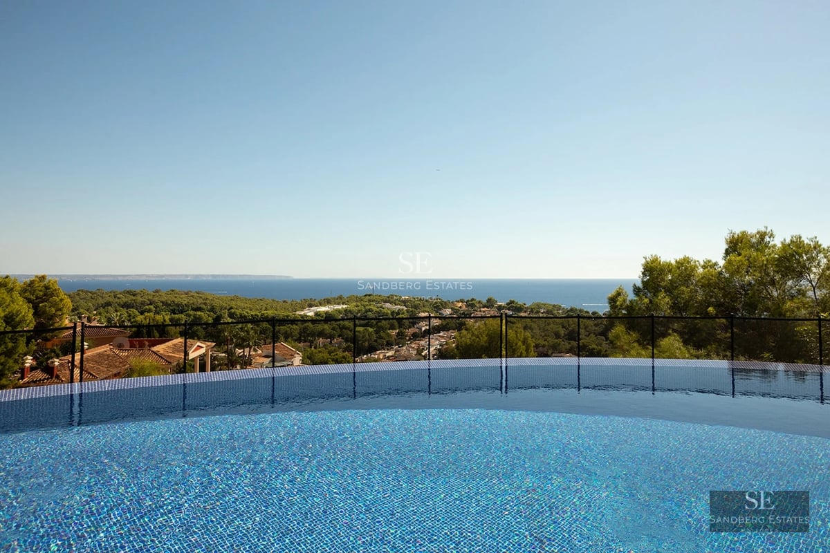 An infinity pool with blue tiles overlooking a lush green forest and the Mediterranean sea under a clear blue sky.