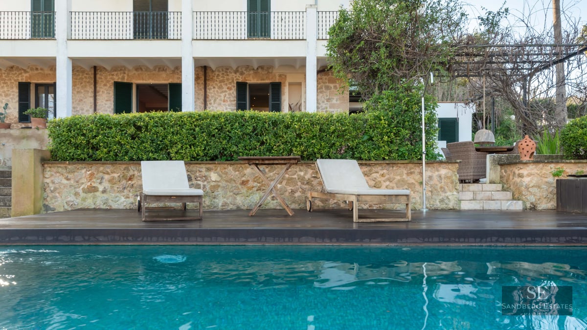 Turquoise swimming pool with wooden loungers in front of a stone Mediterranean house with green shutters.