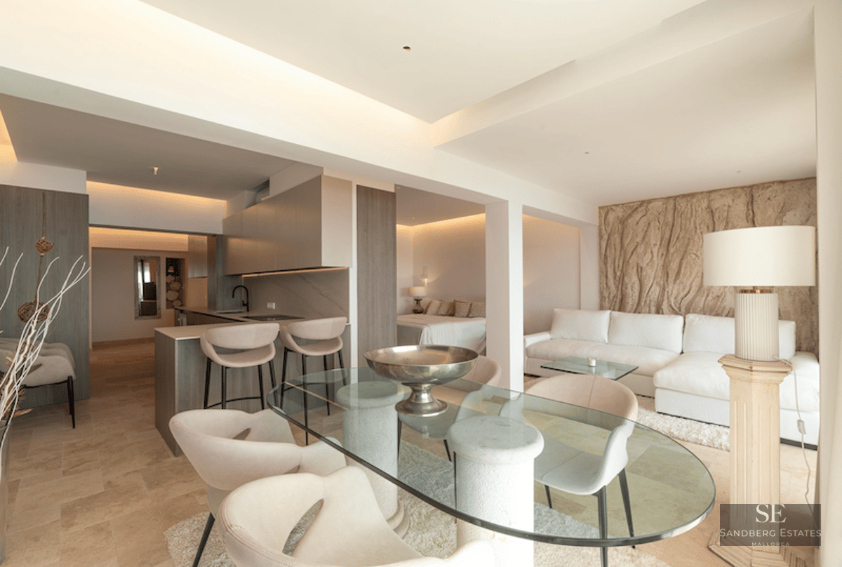 Modern open-plan living room featuring a glass dining table, cream sofa, and integrated kitchen in neutral stone tones.
