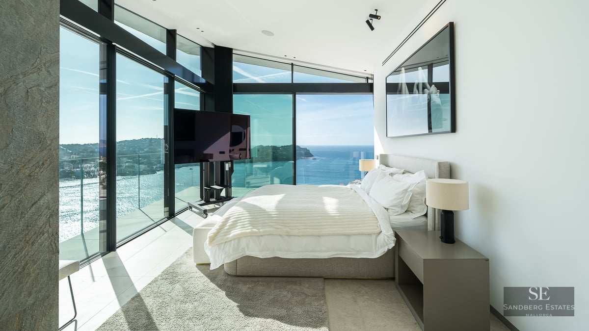 Bright master bedroom with floor-to-ceiling glass windows looking out over the ocean and a coastal town.