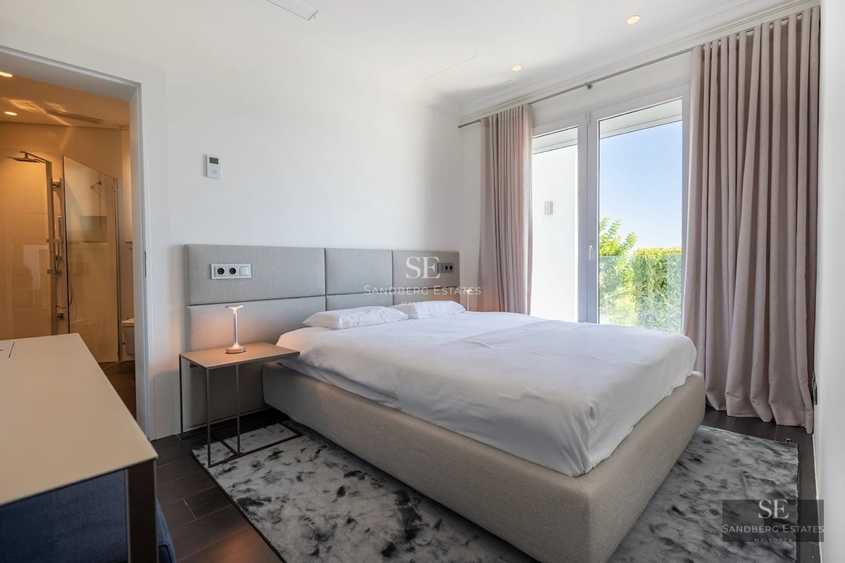 Minimalist bedroom with a large bed, ensuite bathroom view, and bright floor-to-ceiling windows with soft curtains.
