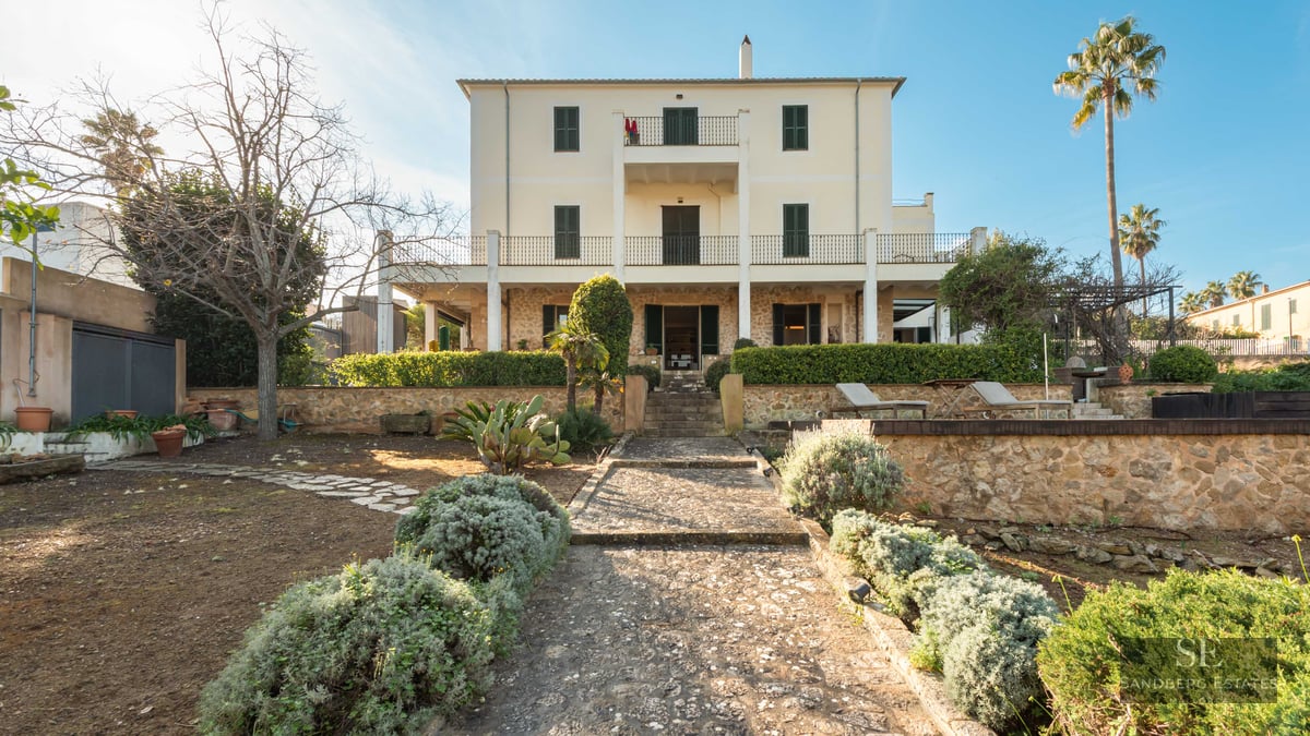 Rear view of a cream three-story villa with stone walls, green shutters, and a stone path through a garden.