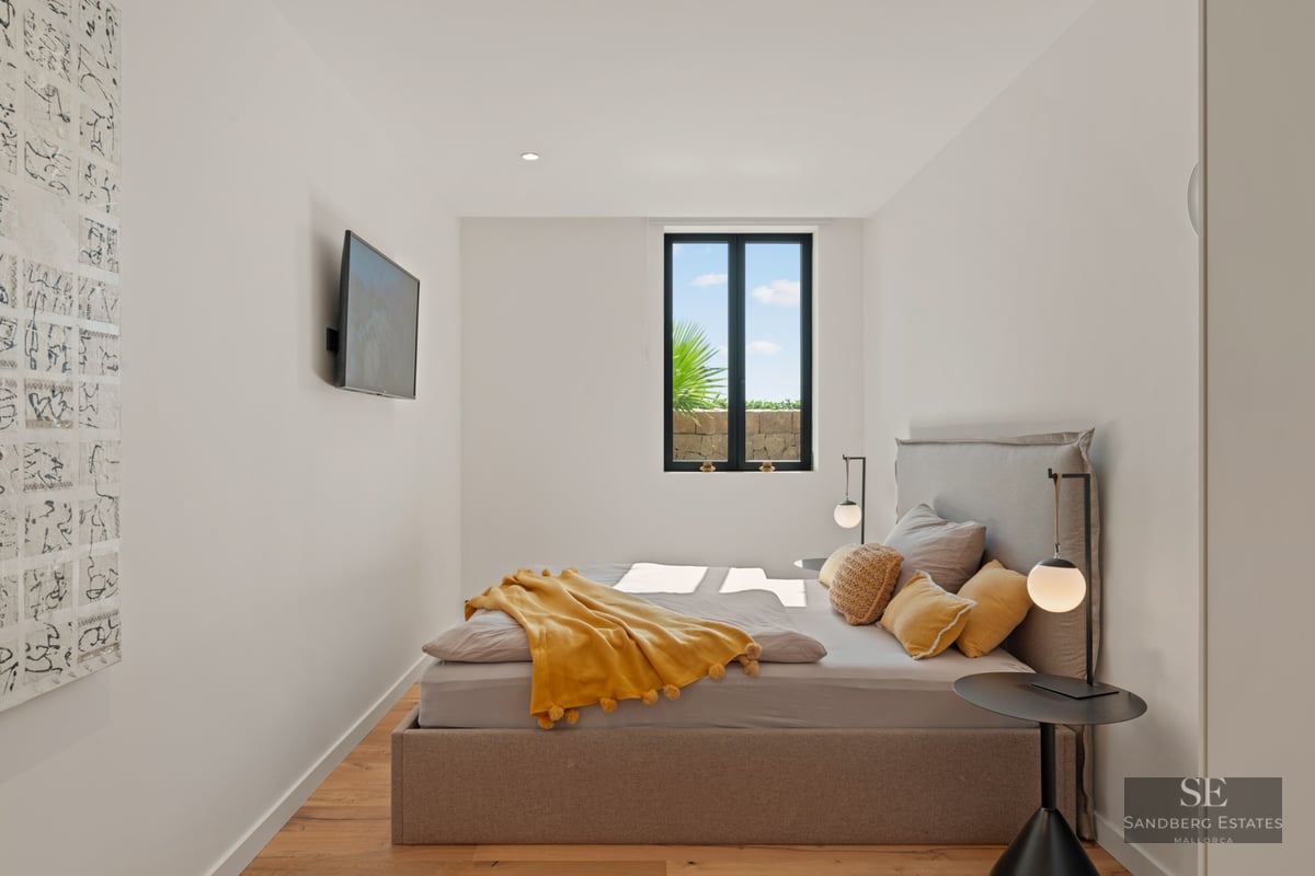 Bright bedroom featuring a taupe upholstered bed, yellow pom-pom blanket, minimalist lamps, and a scenic window view.