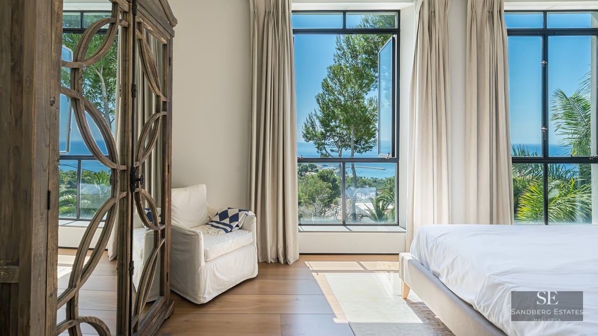 Bright master bedroom with large windows overlooking the sea, featuring a white armchair and ornate wooden wardrobe.