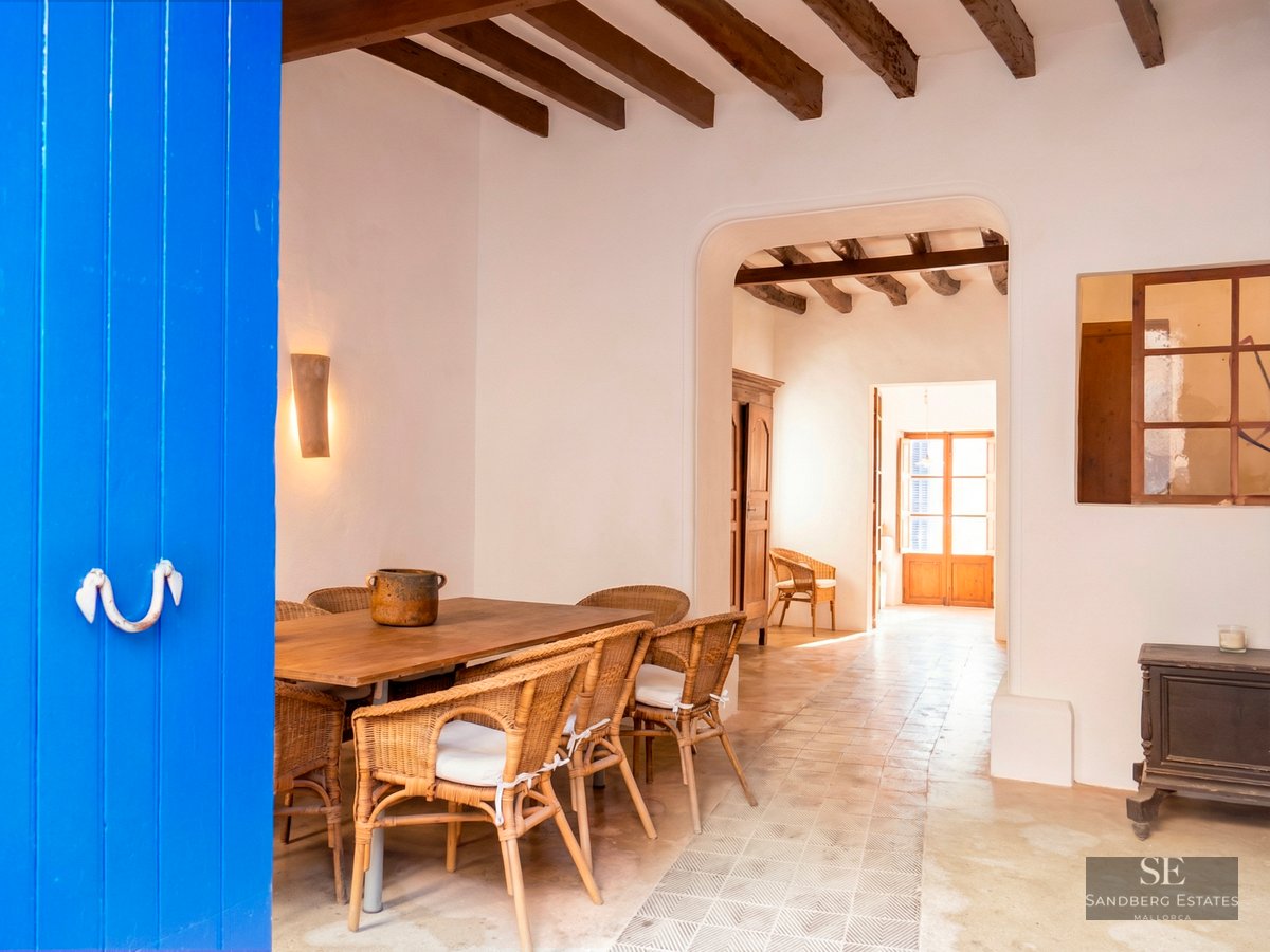 Dining area featuring a wooden table, wicker chairs, exposed ceiling beams, and a striking blue door in the foreground.