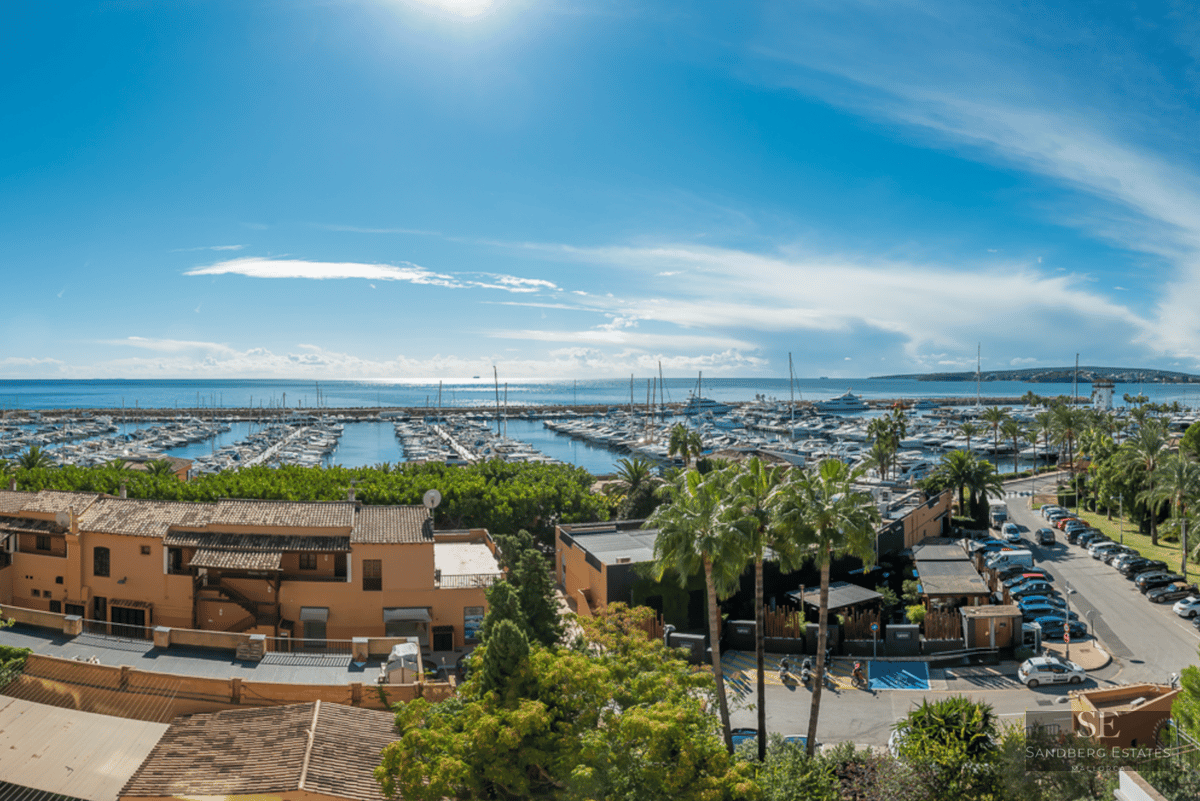 Elevated view of a Mediterranean port with yachts, terracotta buildings, and palm trees under a bright blue sky.