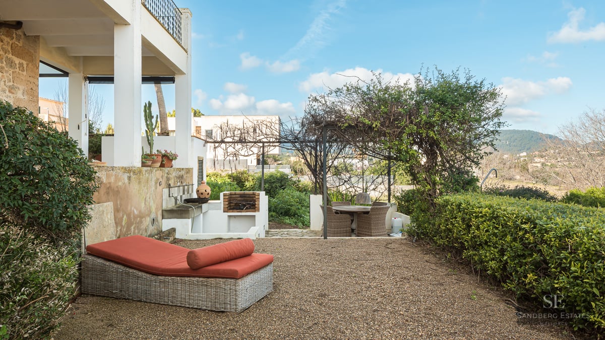 Outdoor terrace featuring a wicker sunbed with orange cushions, a built-in stone grill, and a pergola-shaded dining area.