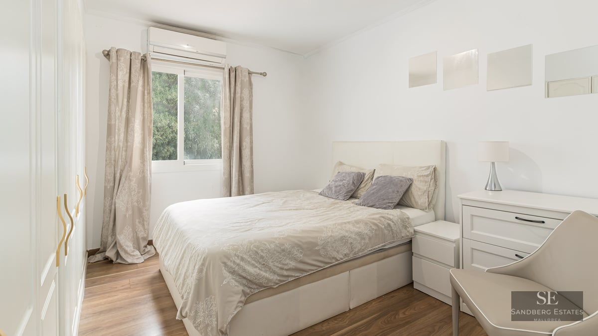 Chambre lumineuse avec lit double, meubles blancs, parquet et fenêtre donnant sur des arbres.