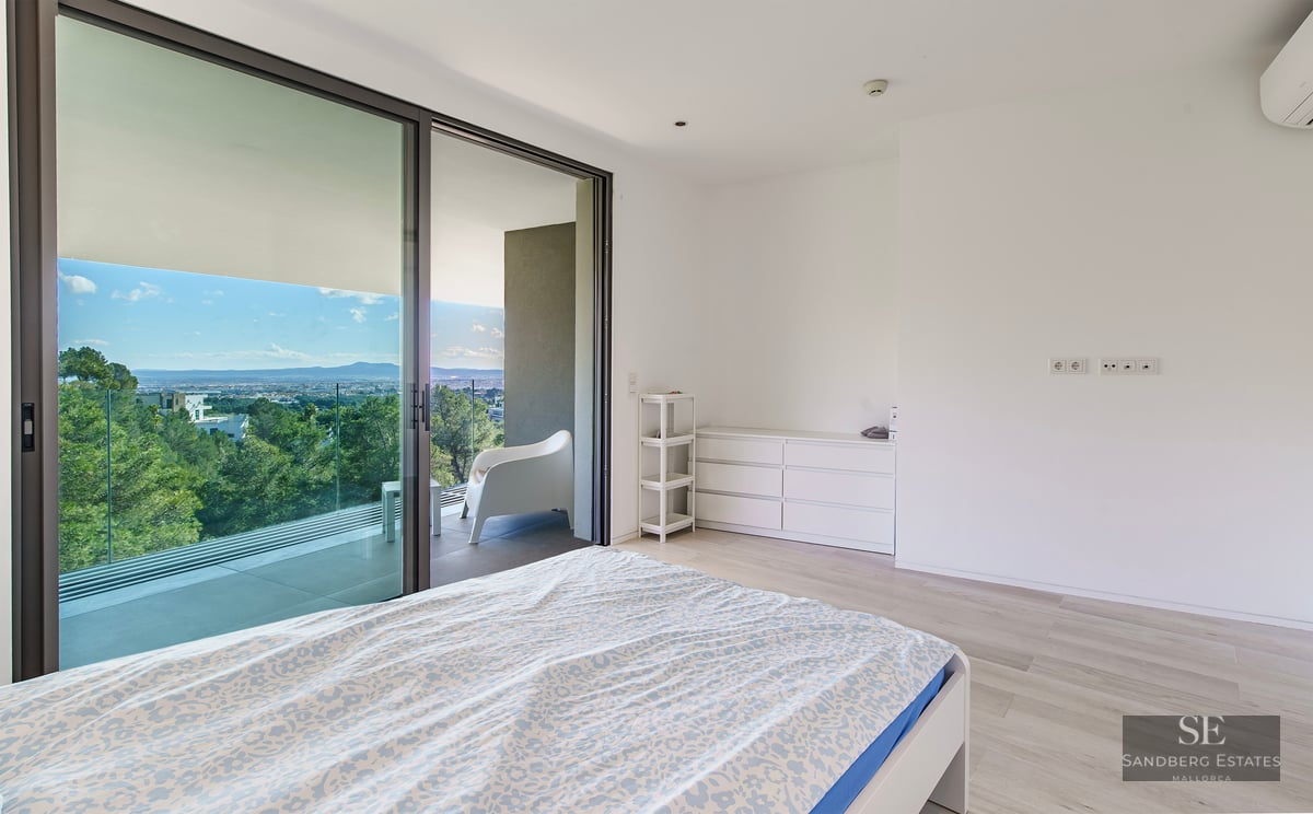 Bright minimalist bedroom with large glass doors opening to a terrace with forest views.