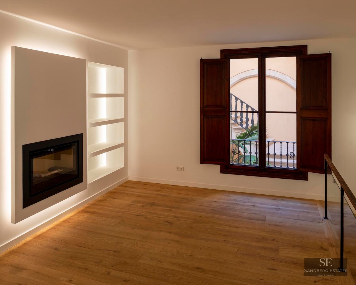 Modern empty room featuring a built-in fireplace, backlit shelving, oak floors, and dark wood shutters looking out to a courtyard.