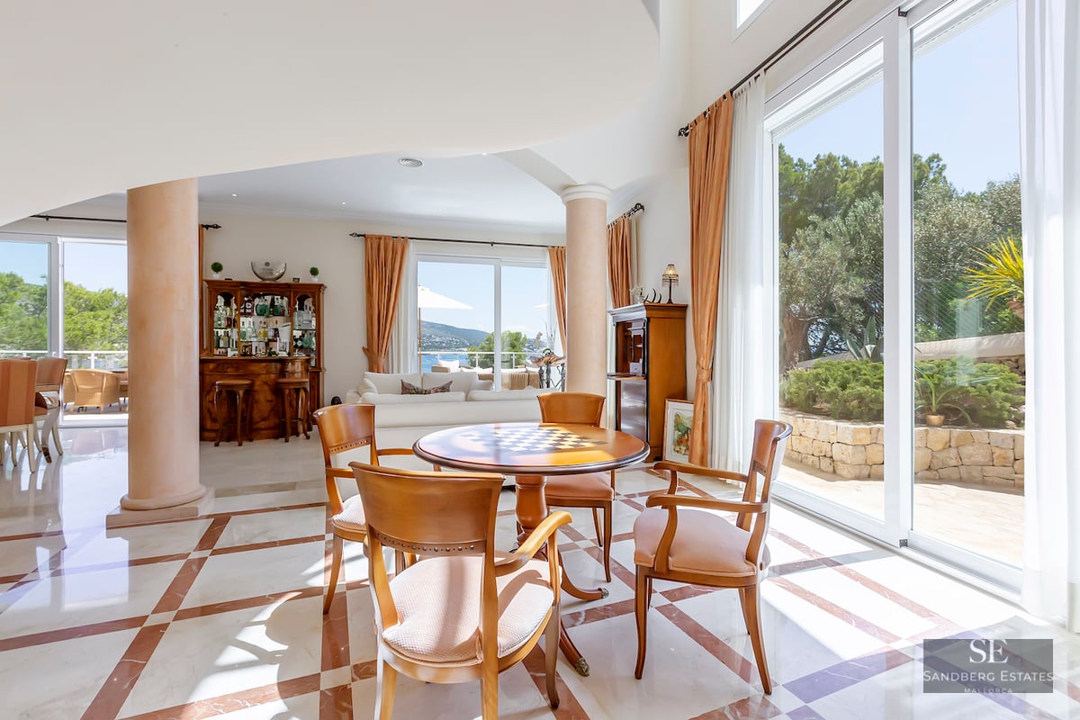 Spacious living area with marble floors, wooden table, pillars, and large windows overlooking a garden and sea.