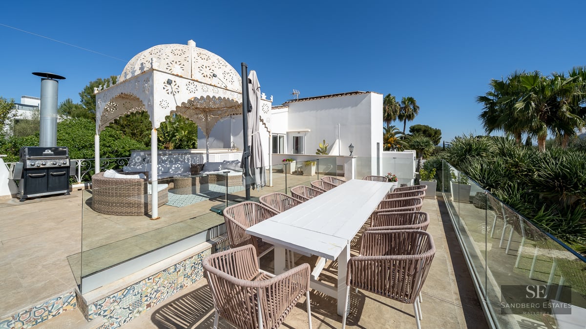 Spacious terrace with a white dining table, wicker chairs, BBQ station, and an ornate white gazebo under a clear blue sky.