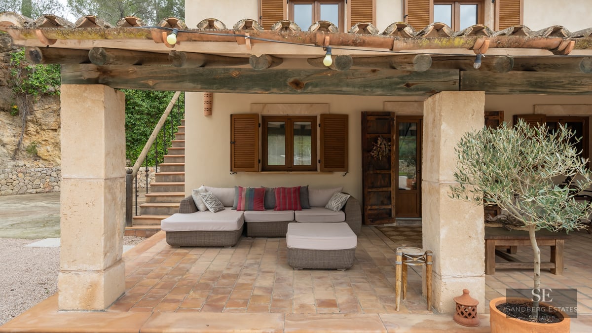 Covered terrace with wooden beams, stone pillars, comfortable sofa, and a potted olive tree.