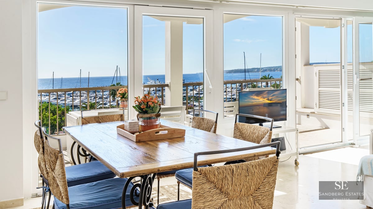 Sunlit dining room with a wooden table and woven chairs looking out over a yacht-filled marina and the blue ocean.