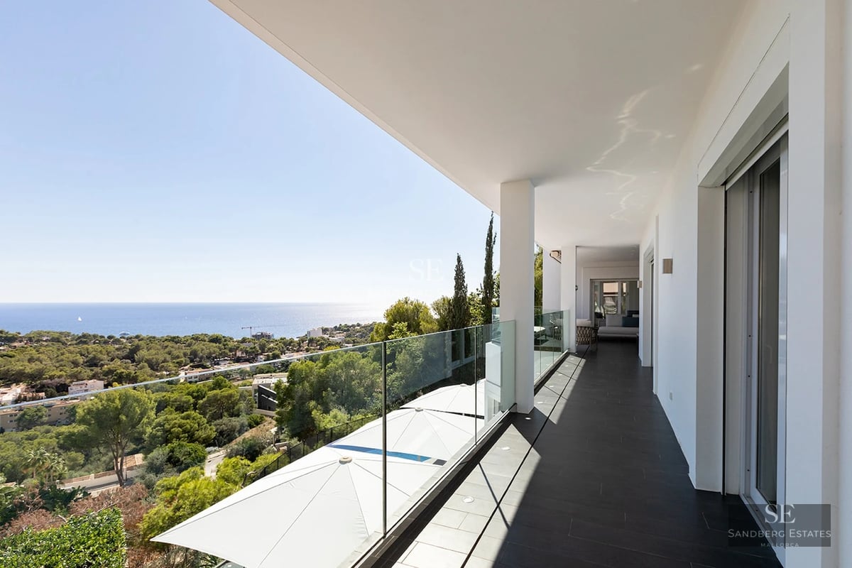 A long white modern balcony with a glass railing overlooking green trees and the blue sea under a clear sky.