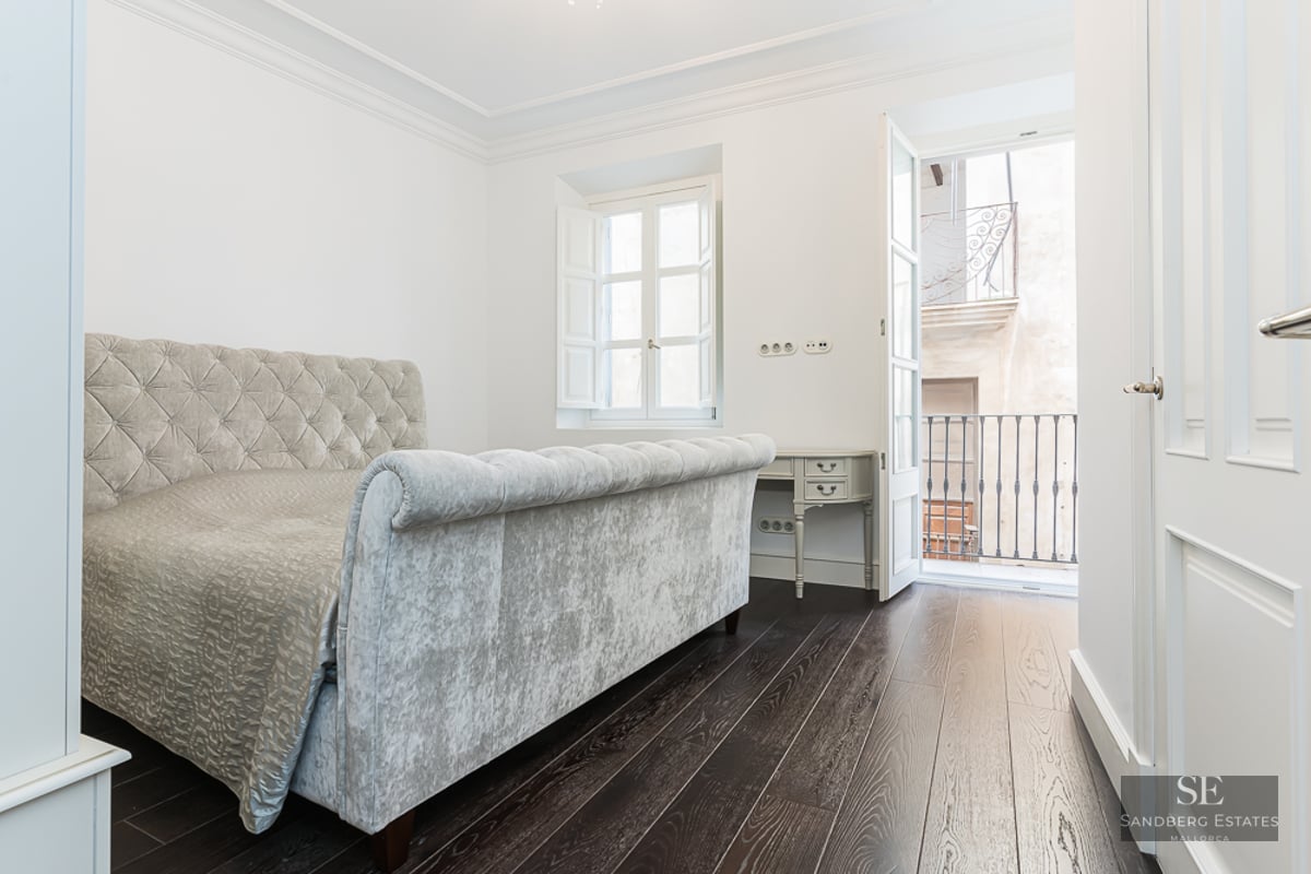 Bright bedroom featuring a grey velvet tufted bed, dark wood floors, and a balcony with wrought iron railing.