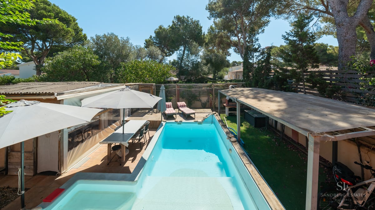 Rectangular turquoise pool surrounded by wooden decking, outdoor dining area, and lush trees under a clear blue sky.