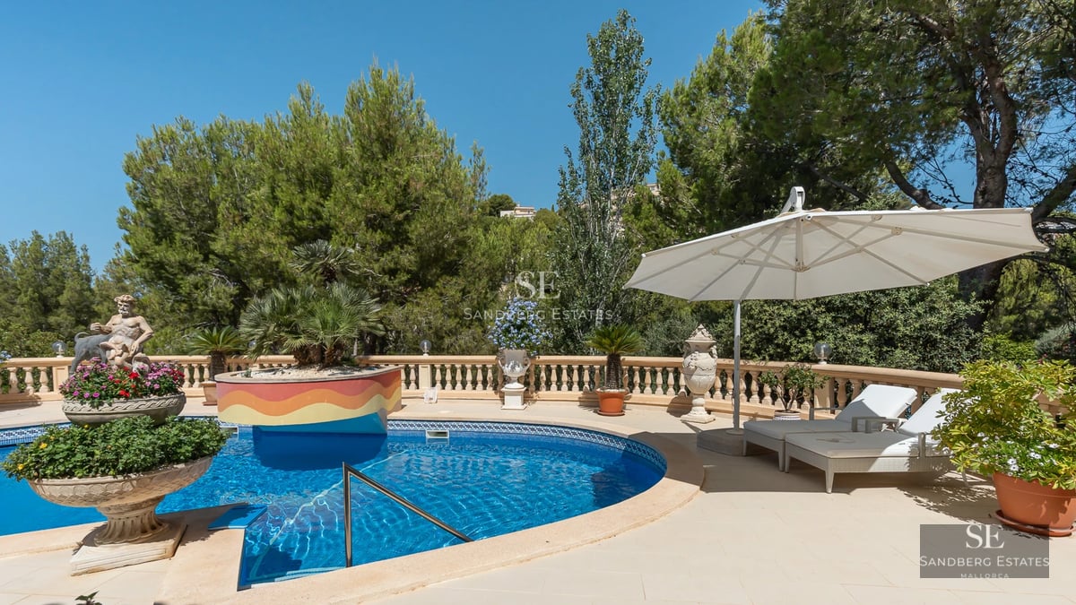Image of a modern villa facade with reflective pool, surrounded by greenery and a bright blue sky.