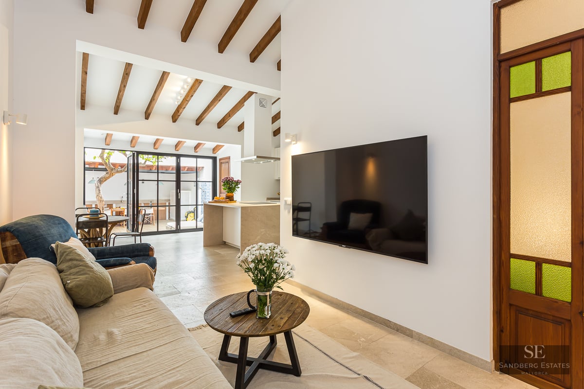 Modern living room featuring a beige sofa, wooden ceiling beams, large flat-screen TV, and a view into an open kitchen.