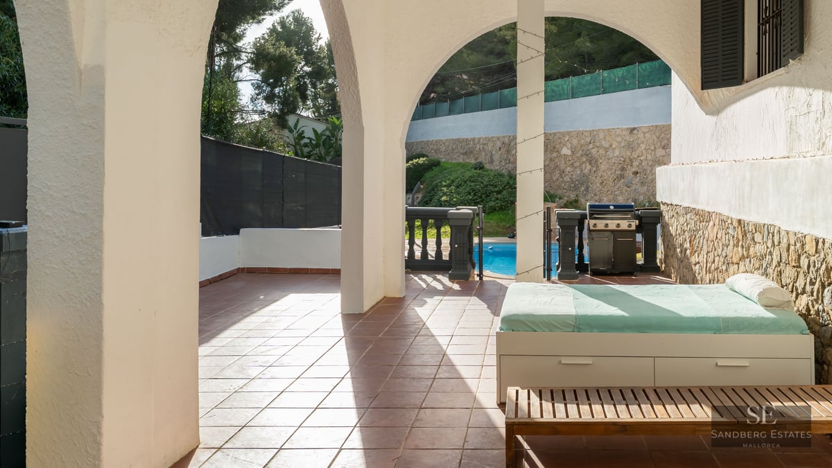 Terrasse couverte avec arches blanches, lit de repos, barbecue et vue sur une piscine ensoleillée.