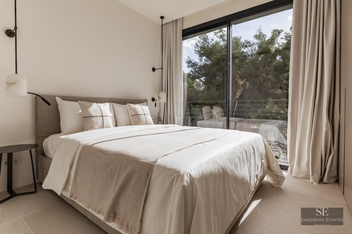 Bright bedroom with neutral-toned bed facing a large floor-to-ceiling window overlooking trees.