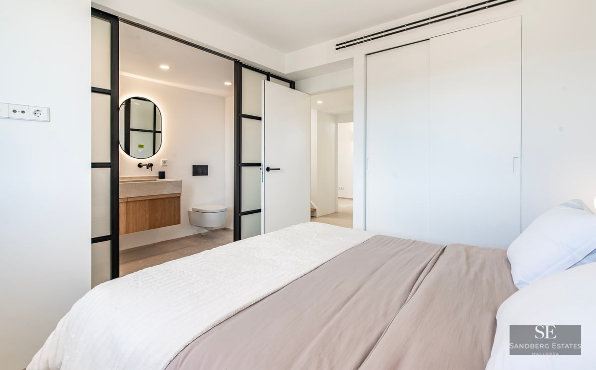 Modern bedroom featuring a bed with neutral linens and a glass-partitioned ensuite bathroom with a backlit round mirror.