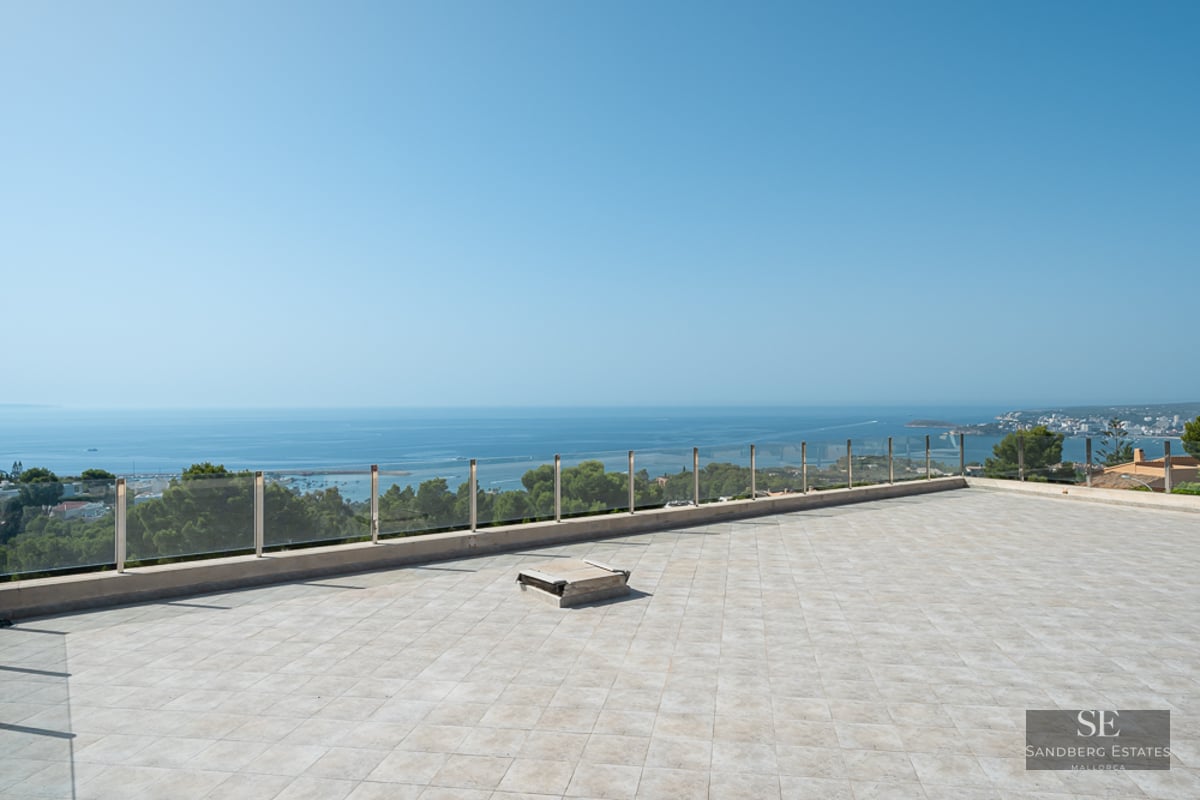 A large stone-tiled terrace with a glass railing overlooking a vast blue ocean under a clear sky.