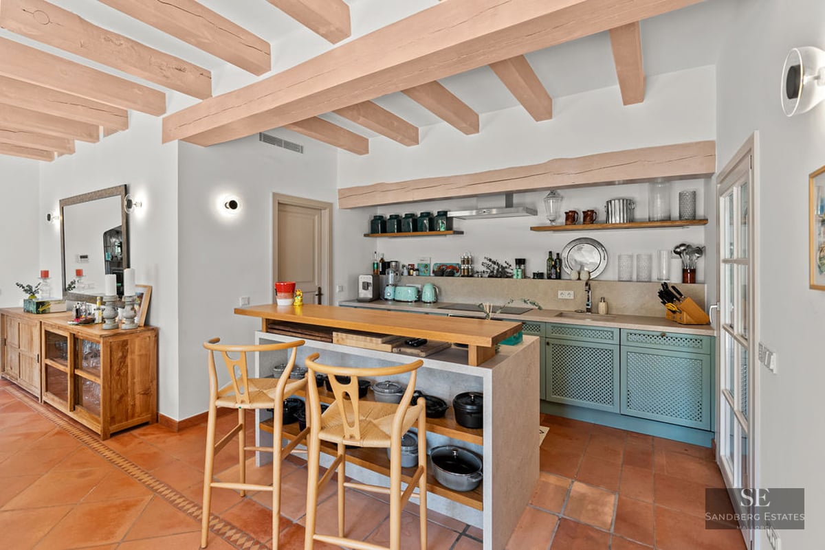 Open-plan kitchen with exposed beams, island with stools, sage green cabinets, and terracotta tile flooring.