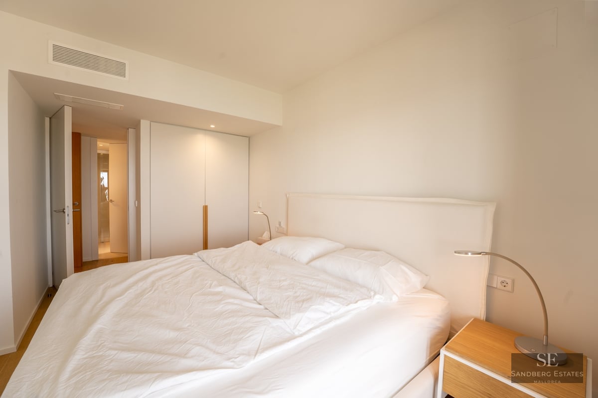 Bright minimalist bedroom with a large bed, white linens, built-in closet, and wooden bedside table.